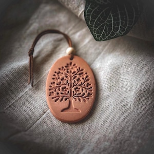 May include: An oval-shaped terracotta ornament featuring an intricate embossed tree design. The piece includes a brown leather cord threaded through a hole at the top, accented by a small, light-colored wooden bead, creating a hanging decoration.