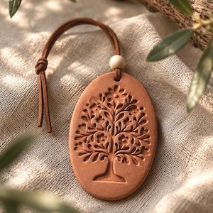 May include: An oval terracotta ornament featuring an embossed tree design with intricate branches and flowers. The piece includes a brown leather cord threaded through a hole at the top, secured by a small, round, light-colored wooden bead.