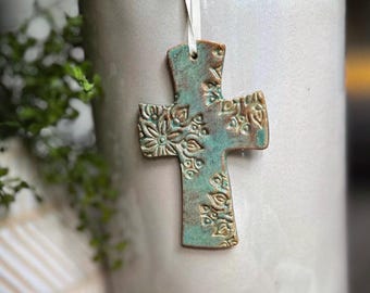 May include: A ceramic cross ornament featuring a textured, embossed floral pattern with a weathered teal and brown glaze finish. The cross is suspended by a thin white ribbon, showcasing intricate botanical details across its entire surface.