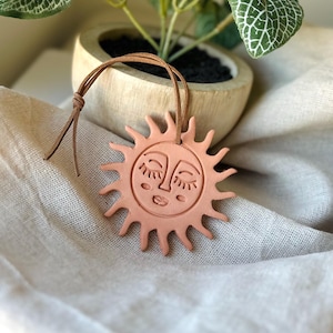 May include: A terracotta sun-shaped ornament with a smiling face and closed eyes. The sun has pointed rays and a brown leather cord for hanging. The ornament is displayed on a neutral-colored fabric, with a potted plant in the background.