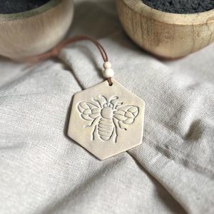 May include: A beige ceramic hexagon-shaped ornament with a white bee design hanging from a brown leather cord with white beads.