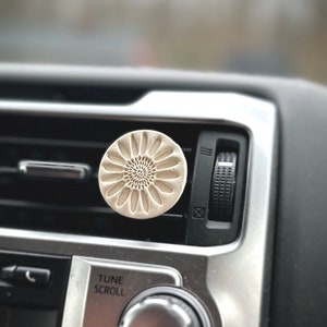 May include: A white ceramic car air freshener with a floral design. The air freshener is attached to the air vent of a car.