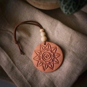 May include: A circular terracotta-colored clay ornament featuring an embossed sun design with a central spiral. The ornament is attached to a brown leather cord accented with three small, round, light-colored wooden beads at the top.