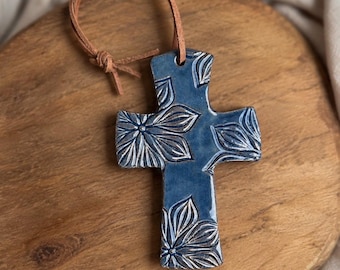 May include: A blue ceramic cross ornament featuring a debossed floral pattern with silver-toned highlights. The cross has a smooth, glossy finish and is suspended by a thin, light brown leather cord tied in a simple knot at the top.