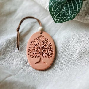 May include: An oval-shaped terracotta-colored clay ornament featuring an embossed tree design. The ornament is suspended by a thin brown leather cord with a small white bead accent at the top, creating a simple and functional hanging decoration.