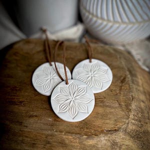May include: Three circular white clay ornaments featuring an embossed floral petal design. Each ornament is suspended by a thin brown leather cord attached through a small hole at the top, creating a uniform set of hanging decorations.