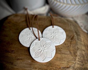 May include: Three circular white clay ornaments featuring an embossed floral petal design. Each ornament is suspended by a thin brown leather cord attached through a small hole at the top, creating a uniform set of hanging decorations.