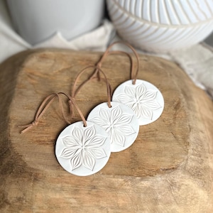 May include: Three circular white ornaments featuring a debossed floral pattern with pointed petals. Each ornament is attached to a thin brown leather cord for hanging. The ornaments have a smooth, matte finish and a consistent, uniform design.