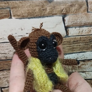 May include: A handmade crochet monkey doll with a brown body, black face, and large black eyes. It wears a yellow and black vest. The doll is held in a person's hand.