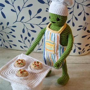 May include: A green crocheted chef doll wearing a white chef's hat and a striped apron. The doll is holding a white crocheted tray with three miniature pizzas. The background features a blue and white floral pattern.