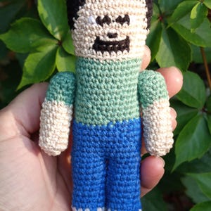 May include: A handmade crochet doll featuring a pixelated design. The doll has a black hair, a tan face, a green shirt, blue pants, and tan arms. The doll is held in a hand against a background of green leaves.