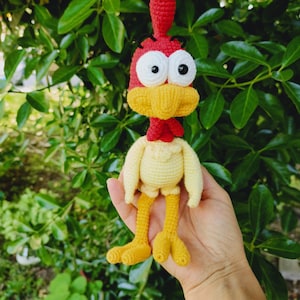 Puede incluir: Un juguete de pollo de crochet con una cresta roja, un cuerpo amarillo y grandes ojos blancos. El pollo está siendo sostenido en la mano de una persona.