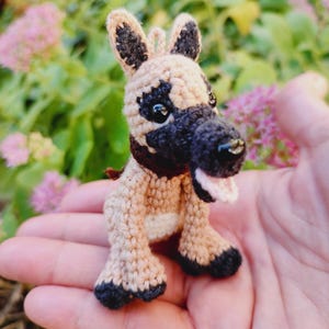 May include: A small crocheted dog toy with brown and black fur, black eyes, and a pink tongue sticking out. The dog is sitting on a hand.
