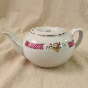 Peut inclure: Théière en céramique blanche avec couvercle et anse. La théière présente une bande à motifs roses et un motif de panier floral. Des bordures dorées ornent le bord et le bec. Une pièce classique pour servir le thé.