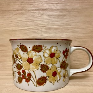 May include: A white ceramic mug with a brown rim and handle. The mug features a floral pattern with brown, yellow, and red flowers.