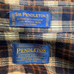 May include: A plaid shirt featuring a brown, navy blue, and tan checkered pattern. Two blue rectangular labels are stitched onto the fabric. The top label reads Sir Pendleton, Assembled in Mexico of Fine Worsted Wool U.S.A Fabric. The bottom label reads Warranted to be a Pendleton, 100% Pure Virgin Wool, Portland, Oregon, Since 1863, M.