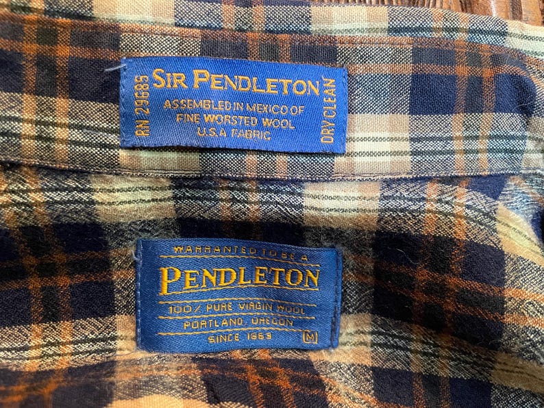 May include: A plaid shirt featuring a brown, navy blue, and tan checkered pattern. Two blue rectangular labels are stitched onto the fabric. The top label reads Sir Pendleton, Assembled in Mexico of Fine Worsted Wool U.S.A Fabric. The bottom label reads Warranted to be a Pendleton, 100% Pure Virgin Wool, Portland, Oregon, Since 1863, M.
