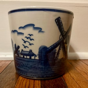 May include: A ceramic planter with a blue and white Dutch windmill design. The cylindrical pot features a landscape with windmills, clouds, and birds. The rim is trimmed with a blue band.