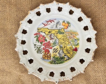 May include: A white ceramic decorative plate with a scalloped, pierced edge and gold trim. The center features a colorful map of Florida, labeled 'Florida The Sunshine State', with illustrations of oranges, flowers, birds, and marine life.
