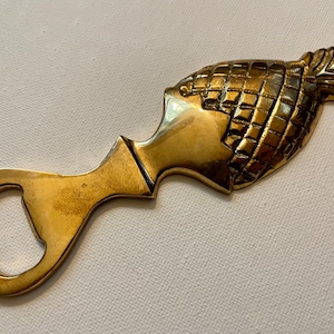 May include: A gold-coloured bottle opener shaped like a pineapple. The opener has a smooth, rounded handle and a bottle opener on the other end.
