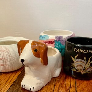 May include: A collection of ceramic planters and a mug. The planters include a seashell, a dog, and a painted design. The black mug has "Cancun" printed on it, along with a seashell and plant design.