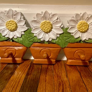 May include: Three wooden daisy flower decorations in orange pots. Each flower has white petals and a yellow center. Green leaves are beneath the flowers. The pots have a wooden knob on the front. The decorations are on a wooden surface.