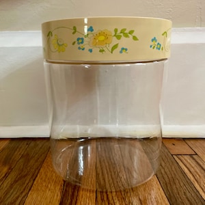 May include: A clear plastic canister with a beige lid. The lid is decorated with a floral pattern in yellow, blue, and green. The canister is sitting on a wooden floor.