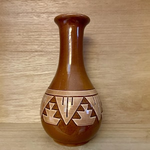 May include: A brown ceramic vase with a narrow neck and a rounded base. The vase features a geometric pattern in a lighter brown colour around the middle. The vase is approximately 18 cm tall.