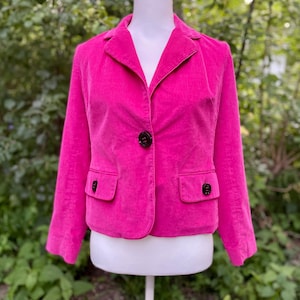 May include: A bright pink corduroy blazer with a notched collar, two front pockets, and a single-button closure. The blazer is displayed on a white mannequin and has dark buttons. The background is a blurred view of green foliage.