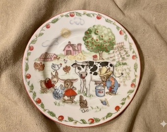 May include: A white ceramic plate with a red rim and a farm scene illustration. The design includes a cow, rabbits, chickens, a barn, and a tree. The plate is decorated with a border of red tomatoes and green leaves.