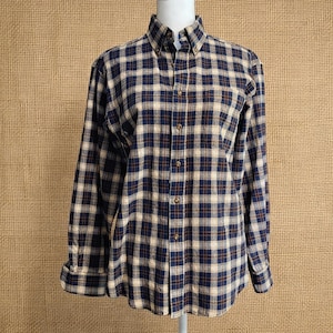 May include: A long-sleeved button-down shirt featuring a plaid pattern in navy blue, tan, and thin dark red lines. The shirt has a pointed collar, a front button placket with matching buttons, and barrel cuffs at the end of each long sleeve.