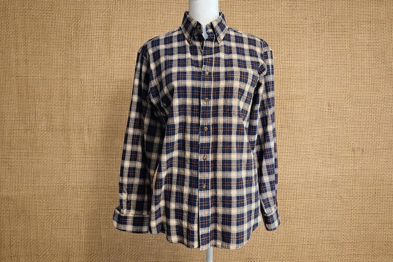 May include: A long-sleeved button-down shirt featuring a plaid pattern in navy blue, tan, and thin dark red lines. The shirt has a pointed collar, a front button placket with matching buttons, and barrel cuffs at the end of each long sleeve.