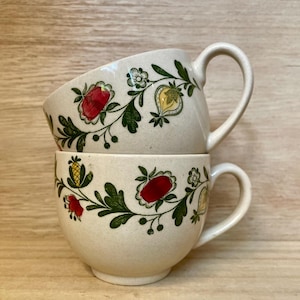 May include: Two stacked cream-colored ceramic mugs with floral designs. Each mug features a repeating pattern of red fruit, yellow fruit, and green leaves. The mugs have curved handles and are set against a light wood background.
