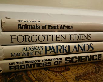 May include: A stack of four books with titles visible: "Animals of East Africa," "Forgotten Edens," "Alaska's Parklands," and "Frontiers of Science." The books have off-white covers with black and blue text. The books are stacked on a patterned surface.