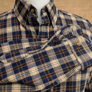 May include: A long-sleeve button-down shirt featuring a plaid pattern in shades of navy blue, tan, and dark brown. The shirt has a pointed collar with visible buttons and a single buttoned cuff on the sleeve, showcasing a textured fabric weave.
