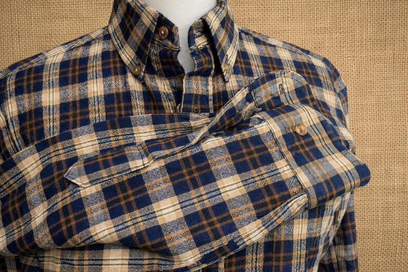May include: A long-sleeve button-down shirt featuring a plaid pattern in shades of navy blue, tan, and dark brown. The shirt has a pointed collar with visible buttons and a single buttoned cuff on the sleeve, showcasing a textured fabric weave.