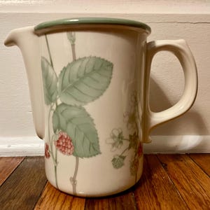 May include: Cream-colored ceramic pitcher with a green rim and handle. The pitcher is decorated with a botanical design of green leaves, red berries, and white flowers. The pitcher has a spout for pouring.