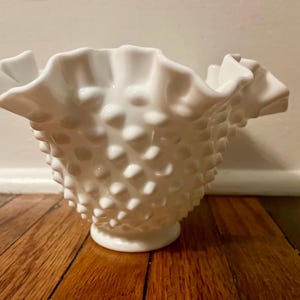 May include: A white hobnail glass bowl with a ruffled rim. The bowl has a textured surface with small, rounded bumps. The bowl sits on a small, circular base. The background is a light-colored wall and a wooden floor.