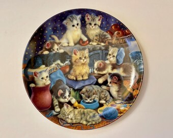 May include: A decorative ceramic plate featuring a whimsical scene of a group of cats in various poses. The plate has a blue background with white stars and features a variety of colors, including brown, white, black, orange, and gray.