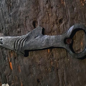 Puede incluir: Un abridor de botellas artesanal con forma de tiburón, hecho de metal oscuro. El abridor presenta una cabeza de tiburón detallada con branquias y una aleta visibles, y un asa circular. El metal tiene un aspecto texturizado y rústico.