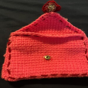 May include: A pink crocheted pouch with red stitching and a gold snap closure. The pouch has a red button on top.