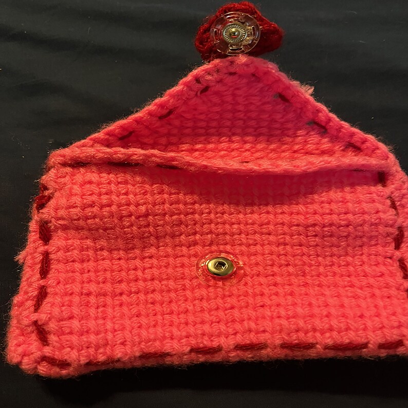 May include: A pink crocheted pouch with red stitching and a gold snap closure. The pouch has a red button on top.