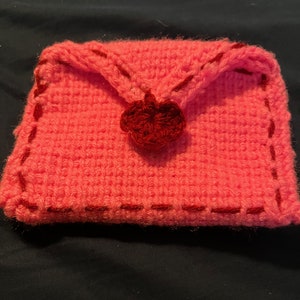 May include: A pink crocheted envelope with a red crocheted heart on the front. The envelope is decorated with red stitching around the edges.