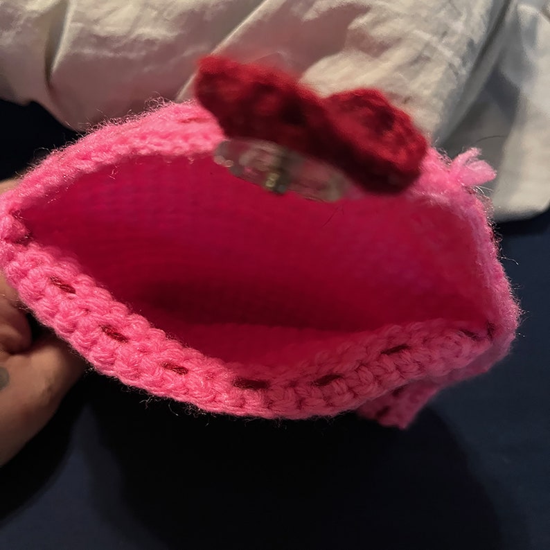 May include: A pink crocheted pouch with a red felt bow on top. The pouch is open and the inside is visible.
