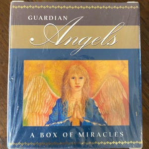 May include: A box of Guardian Angels oracle cards with a blue background and a gold border. The box features a painting of an angel with wings, wearing a blue robe and praying with hands clasped. The text on the box reads "Guardian Angels A Box of Miracles".