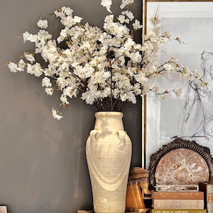 May include: A mirrored cabinet with a white ceramic vase filled with white blossoms. The cabinet has two drawers and two doors with a criss-cross pattern. The cabinet is sitting on a dark brown rug. The cabinet is decorated with books and a framed drawing of a woman.