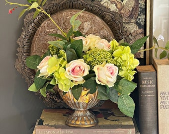 May include: A decorative arrangement featuring a gold-toned vase filled with pink and yellow roses and green hydrangeas. The vase sits atop a stack of books, with a vintage tapestry and a decorative tray in the background. White floral branches add height.