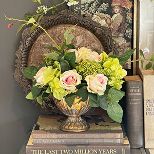 Peut inclure: Composition décorative avec un vase doré rempli de roses roses et jaunes et d'hortensias verts. Le vase est posé sur une pile de livres, avec une tapisserie vintage et un plateau décoratif en arrière-plan. Des branches fleuries blanches ajoutent de la hauteur.