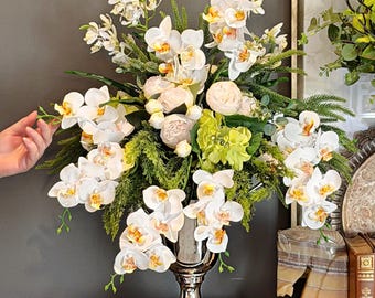 May include: A large floral arrangement featuring white orchids with yellow centers, green foliage, and light pink accents. The arrangement is 24 inches high and 21 inches in diameter, displayed in a silver vase. The text in the image reads: 24" HIGH, 21" DIAMETER.