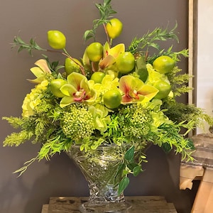 May include: A vibrant floral arrangement in a cut-glass vase. The arrangement features yellow orchids, green lemons, and various green foliage. The vase sits atop a stack of books with the titles "LA RENAISSANCE" and "THE LAST TWO MILLION YEARS".